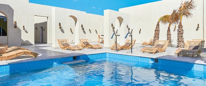 Ein hellblauer Pool liegt sonnig in einem stilvollen Innenhof des True Beach Village. Weiße Wände und moderne Poolmöbel umgeben das Wasser.