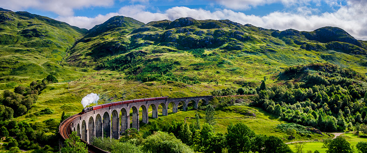 Der Jacobite Steam Train fährt über das historische Glenfinnan Viadukt. Dahinter erstreckt sich die grüne Berglandschaft der schottischen Highlands.